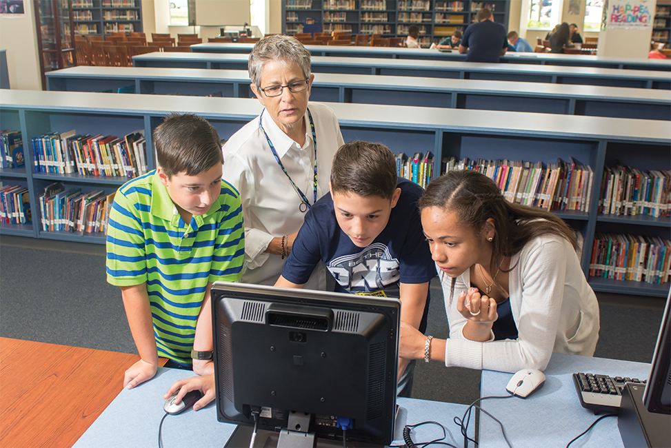 Students doing computer work while the teacher looks on. Students doing computer work while the teacher looks on.